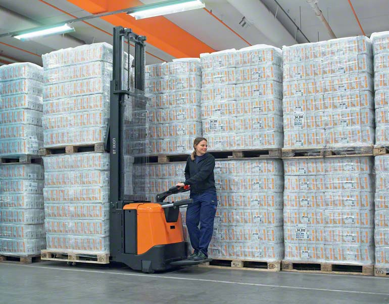 Operator stojący na podeście wózka paletowego elektrycznego w trakcie przenoszenia towaru w magazynie